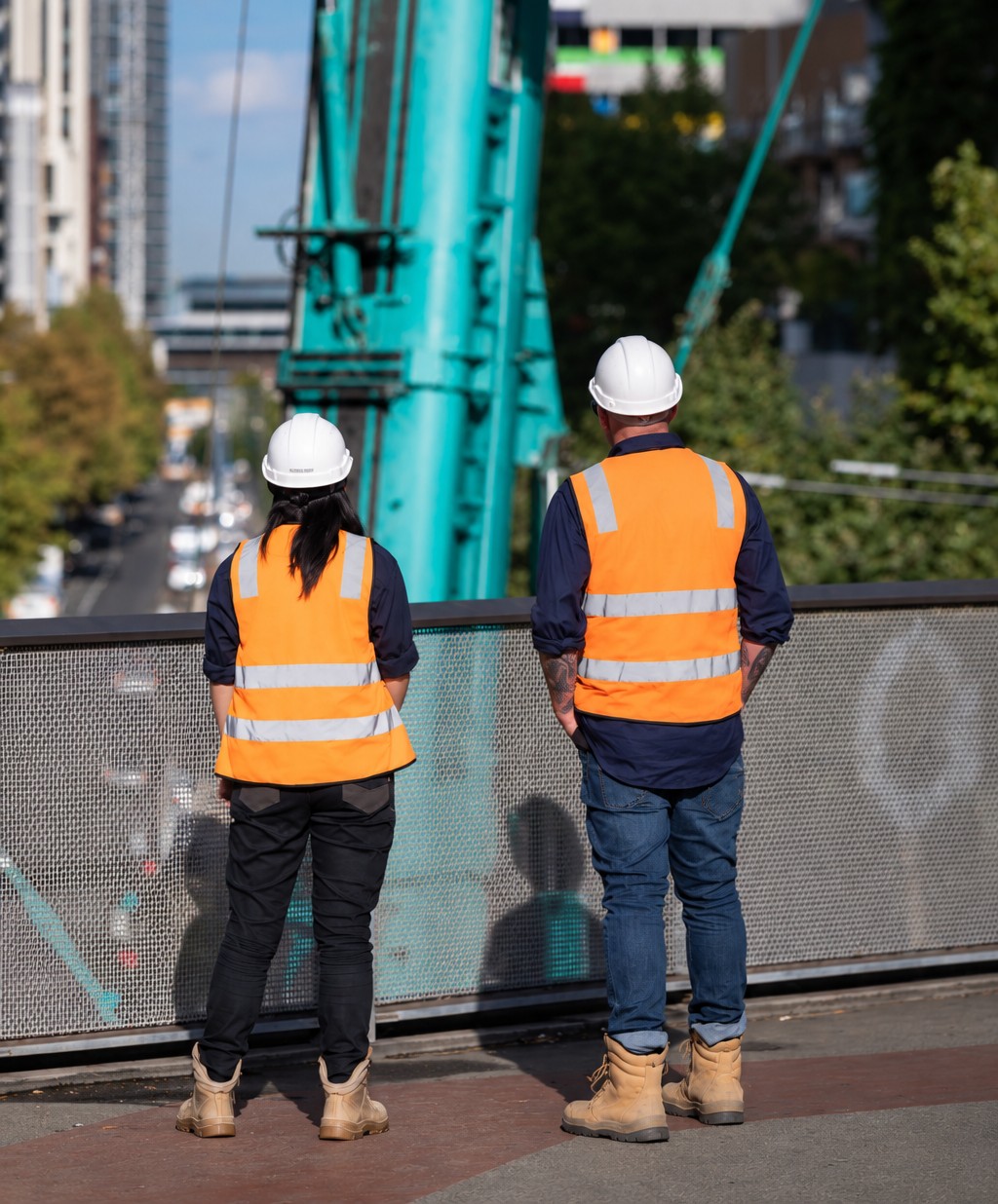 Two Apex workers in high-vis overlooking a city worksite beside a teal crane